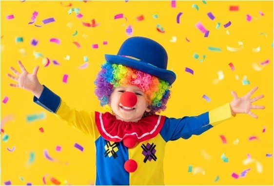 Clown en costume coloré avec perruque et nez rouge, souriant sous des confettis sur fond jaune.