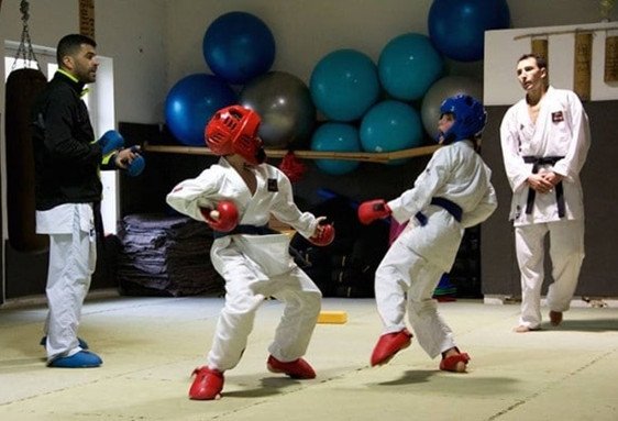 Deux enfants en kimono s’entraînent au karaté sous la supervision de deux coachs dans un dojo.