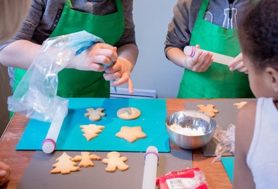 Deux personnes en tabliers verts préparent des biscuits en forme d'étoiles et d'arbres sur une table, avec des emporte-pièces