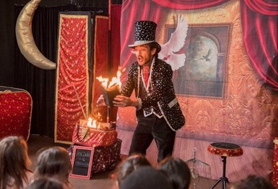 Un magicien en costume noir à pois et haut de forme fait un numéro de feu pour un jeune public.