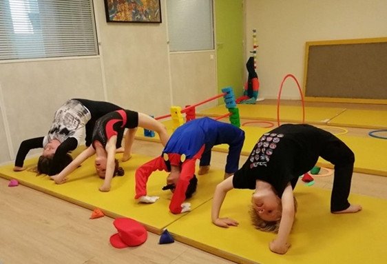 Quatre enfants en cours de danse signent des positions inversées sur un tapis jaune, sous supervision dans une salle dédiée aux arts du mouvement.